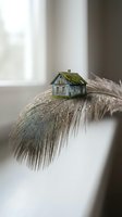 Miniature wooden house with moss roof balanced on delicate feather creating surreal conceptual art about home dreams and lightness.