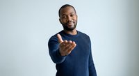 Young African American man in blue sweater making welcoming gesture with open hand on light background for business presentations.