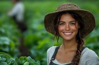 Young Hispanic woman farmer in straw hat and overalls smiling while working in organic vegetable garden, with green plants and natural background.