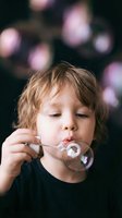 Young blonde boy blowing soap bubbles with concentrated expression against dark background with colorful bokeh lights for childhood and play concepts.