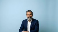 Middle-aged man with salt-and-pepper beard, South Asian ethnicity, wearing navy blazer and white shirt against blue background. Corporate professional portrait for business, leadership, and executive use.