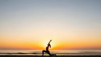 Woman practicing yoga warrior pose on beach at sunrise with golden sky and ocean waves creating peaceful meditation atmosphere for wellness.