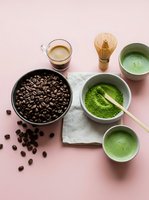 Coffee beans and matcha powder with traditional tea ceremony tools on pink background for beverage preparation and wellness lifestyle concepts.