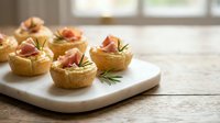 Gourmet appetizer tartlets filled with creamy filling and topped with prosciutto ham on white serving board. Elegant party food for catering events.