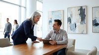 Senior businesswoman mentoring young male colleague in modern office meeting room with abstract art on walls and natural lighting.