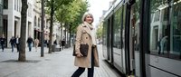 Mature blonde businesswoman in beige coat standing confidently on urban street with modern tram, representing professional success and city commuting lifestyle.