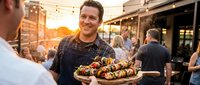 Smiling male chef in apron presenting gourmet appetizer platter at outdoor restaurant during golden hour sunset with customers dining in background.