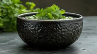 Fresh cilantro leaves in textured black ceramic bowl on dark stone surface, moody food photography with selective focus and natural lighting.