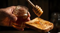 Closeup of hand drizzling golden honey from wooden dipper onto toasted bread slice against dark background, ideal for food and culinary content.