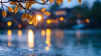 Autumn leaves over calm water surface at sunset with golden bokeh lights reflecting. Moody evening atmosphere with blurred background creates peaceful nature scene.