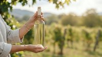 Person holding bottle of white wine with vineyard landscape in background, suitable for wine industry, agriculture, and beverage marketing.