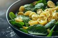 Fresh tortellini pasta with baby spinach leaves in dark ceramic bowl on rustic background, macro food photography with selective focus and natural lighting.