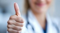 Closeup of medical professional showing thumbs up gesture wearing white coat, expressing positivity and success in healthcare setting. Blurred background.