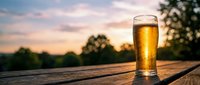 Golden beer glass on wooden table during sunset with blurred trees and colorful sky background creating peaceful outdoor drinking atmosphere.