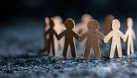 Paper cutout figures holding hands in unity chain against dark background, symbolizing community, teamwork and social connection. Shallow depth of field creates dramatic mood.