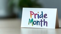 Rainbow colored lettering spelling Pride Month on white greeting card against blurred green background. Text features cheerful watercolor style with vibrant gradient hues.