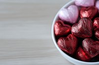 Red and pink foil wrapped chocolate hearts in white ceramic bowl on wooden surface, romantic holiday candy composition with copy space for Valentine's Day designs.
