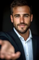 Confident Caucasian businessman in dark suit and white shirt smiling at camera while reaching forward, dramatic lighting creates professional atmosphere.