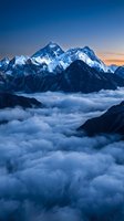 Majestic snow-capped mountain peaks rising above sea of clouds at dawn with dramatic blue and orange sky creating breathtaking alpine landscape scenery.