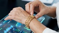 Senior woman with age spots and wrinkles wearing ornate gold bracelet, touching wrist while working at computer with digital interface display, representing technology adoption for older adults.