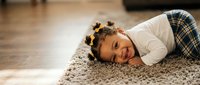 Young African American girl with curly hair lying on soft carpet smiling joyfully in cozy home interior for family lifestyle and childhood happiness concepts.