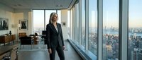 Professional businesswoman in elegant suit standing by floor-to-ceiling windows in modern corporate office with city skyline view during golden hour.