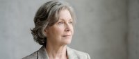 Mature woman with gray hair in updo style, fair complexion, wearing neutral blazer, looking thoughtfully to side against blurred background for corporate or professional use.