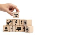 Hand placing wooden cube with biohazard symbol on stack of blocks featuring various hazard and energy icons on white background.