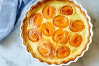 Fresh apricot custard tart in white ceramic baking dish on marble surface, golden fruit arranged in creamy filling, homemade French pastry dessert.