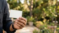 Close-up of mature man's hand holding blank white business card outdoors in garden setting with blurred green foliage background.