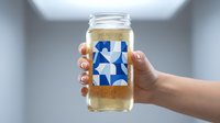 Hand holding glass jar with blue geometric pattern label containing golden liquid in modern kitchen setting for beverage branding and packaging design.