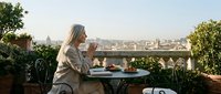 Young blonde woman enjoying morning coffee on rooftop terrace with city skyline view, relaxing outdoor lifestyle moment for wellness and urban living concepts.