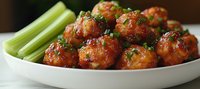 Glazed spicy meatballs garnished with fresh herbs served on white plate with celery sticks, appetizing party food photographed in warm lighting.
