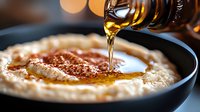 Creamy hummus in dark bowl with olive oil being drizzled from bottle, close-up view with bokeh lights in background. Middle Eastern appetizer preparation.