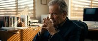 Senior Caucasian businessman in contemplative pose at modern office desk with warm lighting and venetian blinds creating atmospheric shadows.