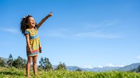 Young girl with curly dark hair, wearing yellow patterned dress, pointing upward while standing in green meadow with mountain landscape and blue sky background.