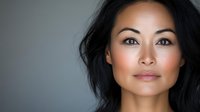 Young Asian woman with natural makeup and serene expression against gray background, professional headshot style portrait showcasing clean beauty and confidence.