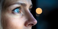 Close-up profile of young woman with bright blue eyes gazing at glowing orange light against dark background, creating mysterious atmospheric mood.