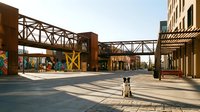 Industrial urban plaza with elevated steel bridges and rustic brick structures casting dramatic shadows on pavement, featuring modern architectural design for contemporary city development projects.