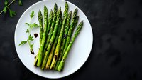 Fresh green asparagus spears arranged on white ceramic plate with herbs and balsamic reduction against dark background, dramatic food photography styling.