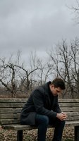 Thoughtful young Caucasian man in dark coat sitting alone on wooden bench in autumn park with bare trees, contemplating life decisions and mental health.