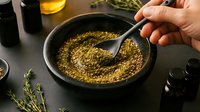 Hand stirring golden mustard seeds in dark bowl with spoon, cooking preparation with herbs and oil bottles on kitchen counter for culinary recipes.