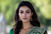 Young South Asian woman wearing traditional green and gold sari with ornate gold earrings, long dark hair, and natural makeup against blurred outdoor background.
