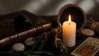 Mystical fortune telling setup with burning candle, tarot cards, crystal ball, dried herbs and ancient books creating atmospheric divination scene.
