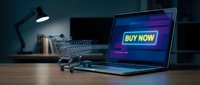 Laptop with glowing buy now button on screen beside a shopping cart on a wooden desk, representing online shopping and e-commerce concepts.