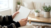Woman in cozy sweater reading blank book in bright living room with plants and natural light for relaxation and leisure activities.