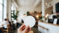 Hand holding blank white circle badge in modern office workspace with blurred colleagues and bright interior background for branding mockup.