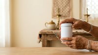 Senior woman with dark skin tone holding white ceramic mug in hands with bracelets, displaying product in warm home interior with religious decor and natural lighting.