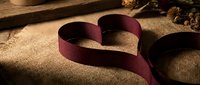 Burgundy ribbon twisted into heart shape on rustic burlap fabric with dried flowers and wooden elements creating romantic Valentine's Day decoration concept.