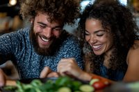 Happy mixed race couple laughing while cooking together in modern kitchen preparing healthy meal with fresh vegetables for romantic dinner date.